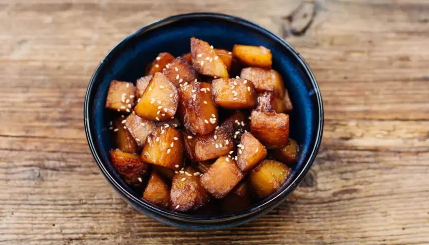 Glasierte Süßkartoffelwürfel mit Sesam, ein köstliches Beispiel für Banchan Rezepte, serviert in einer blauen Schüssel auf Holzuntergrund.