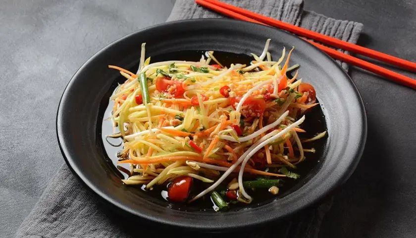 Ein bunter Papaya Salat mit Karottenstreifen, Kirschtomaten und Sprossen, serviert auf einem schwarzen Teller mit roten Stäbchen.
