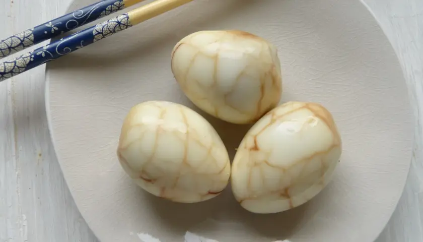 Drei marmorierte Tee Eier auf einer hellen Platte, daneben blaue Essstäbchen.