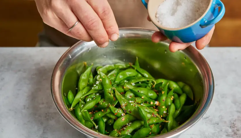 Hände würzen frisch gekochte Edamame mit Meersalz und Chiliflocken. So einfach ist es, Edamame zuzubereiten!