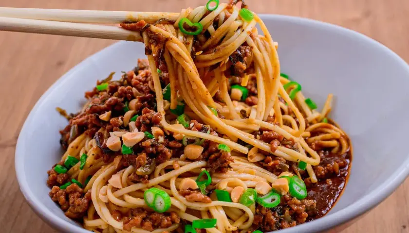 Ein Teller Dan Dan Noodles mit Hackfleisch, Erdnüssen und Frühlingszwiebeln, serviert mit Stäbchen.