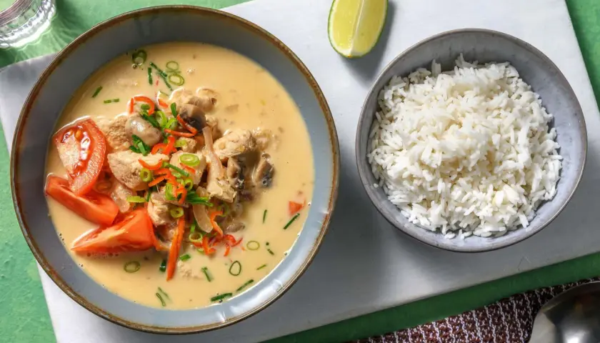 Khao tom: Eine Schüssel mit cremigem Hühner-Kokos-Eintopf, garniert mit Tomaten und Frühlingszwiebeln, daneben eine Schüssel Reis und eine Limettenspalte.
