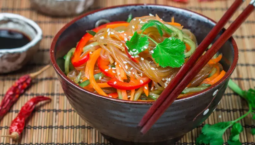 Schüssel mit Japchae, einem koreanischen Nudelgericht, garniert mit Koriander und Sesam. Daneben Sojasauce, getrocknete Chilis und Reis.