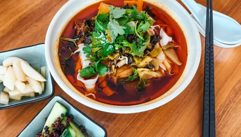 Eine Schüssel Niu Rou Mian, garniert mit Koriander, daneben eingelegte Gurken und Rettich.