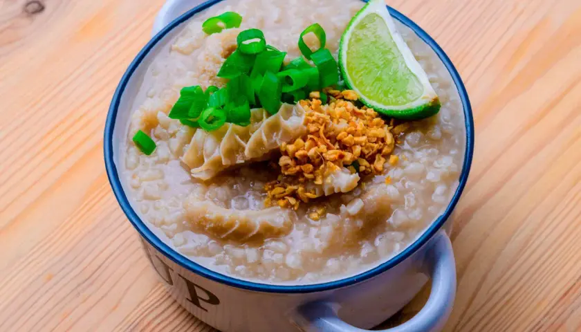 Herzhafter Reis Congee mit Rinderpansen, Frühlingszwiebeln, geröstetem Knoblauch und Limettenspalte in einer Schüssel auf Holzhintergrund.