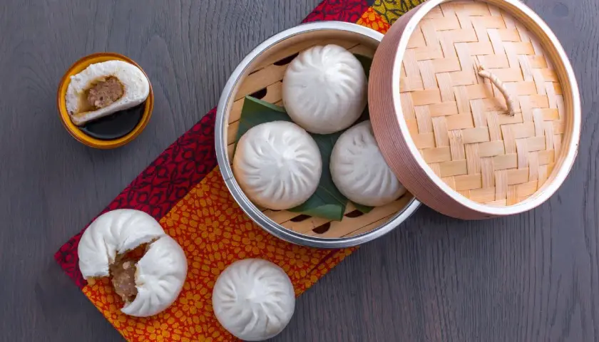 Dampfende chinesische Brote, gefüllt mit herzhaftem Fleisch, werden in einem Bambuskorb serviert. Eines ist aufgeschnitten und zeigt die köstliche Füllung.