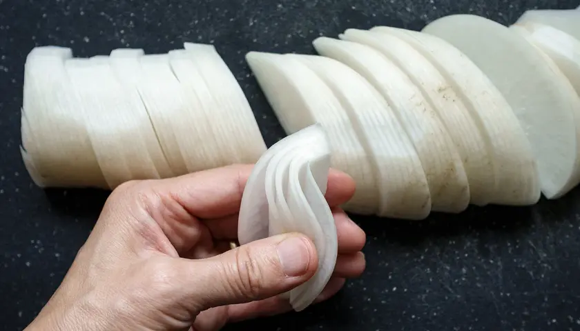 Eine Hand hält dünn geschnittene Rettichscheiben, bereit zum Einlegen. Weitere Scheiben liegen auf einem dunklen Untergrund.