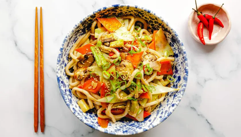 Schüssel mit leckeren Udon Nudeln kochen, garniert mit Gemüse, Frühlingszwiebeln und Sesam. Daneben liegen Essstäbchen und eine kleine Schale mit Chilis.