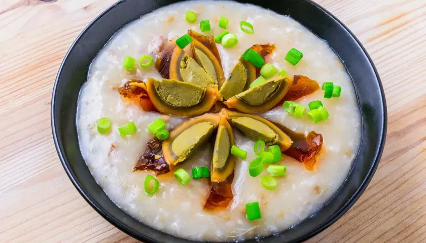 Ein cremiges Congee-Rezept mit geschnittenem konserviertem Ei und Frühlingszwiebeln, serviert in einer schwarzen Schüssel auf Holzhintergrund.