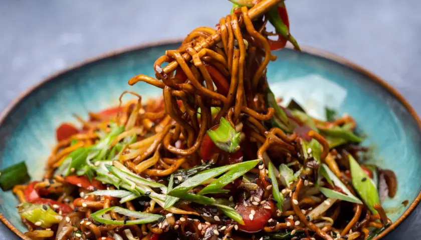 Ein Teller mit dampfendem Chow Mein, garniert mit Frühlingszwiebeln und Sesam. Essstäbchen heben eine Portion der Nudeln an.