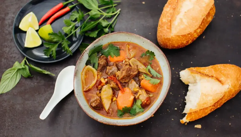 Herzhaftes Bo Kho, ein vietnamesischer Rindfleischeintopf mit Karotten und Sternanis, serviert mit Baguette und frischen Kräutern.