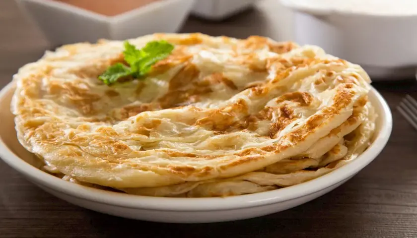 Ein Stapel knuspriger Roti Canai, garniert mit Minze, serviert auf einem Teller mit Curry und Joghurt im Hintergrund.