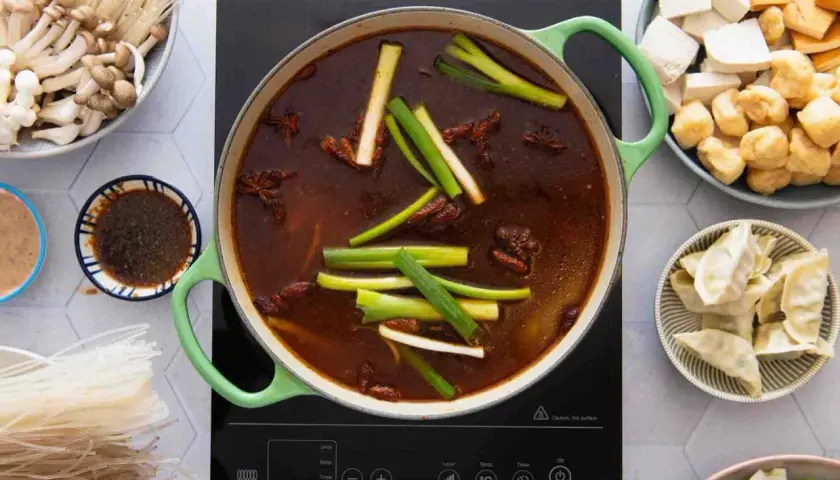 Ein köstlicher chinesischer Eintopf brodelt in einem grünen Topf, umgeben von Zutaten wie Pilzen, Tofu, Teigtaschen und Reisnudeln.