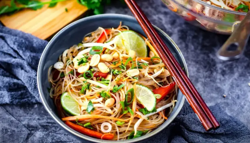 Schüssel mit asiatischem Glasnudelsalat, garniert mit Erdnüssen, Limettenspalten und Frühlingszwiebeln. Chopsticks liegen auf dem Teller.