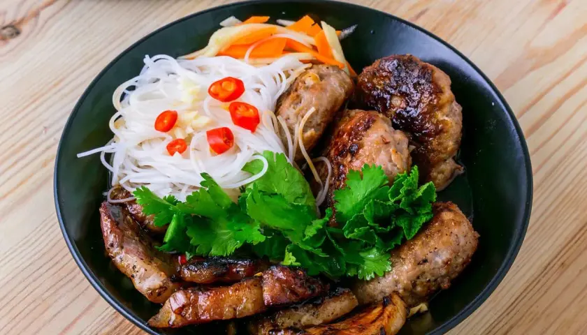 Bun cha Hanoi: gegrilltes Schweinefleisch und Fleischbällchen mit Reisnudeln, Kräutern und Chili auf einem Teller.