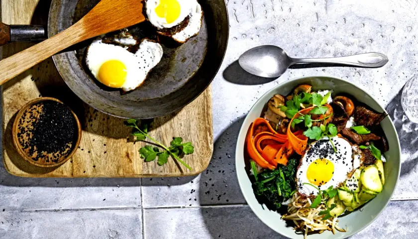 Ein köstliches Bibimbap-Rezept: Bunte Schüssel mit Reis, Gemüse, Rindfleisch, Pilzen und Spiegelei, garniert mit schwarzen Sesamkörnern.