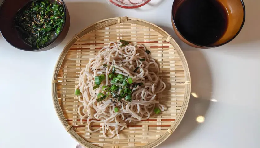 Kalte Soba-Nudeln, garniert mit Frühlingszwiebeln und Nori, serviert auf einem Bambusteller mit Beilagen wie Seetangsalat und Radieschen.