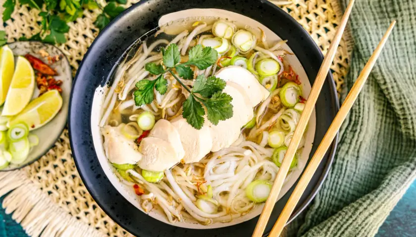 Eine dampfende Schüssel Pho Ga mit zarten Hühnerbrustscheiben, Reisnudeln, Sojasprossen und Frühlingszwiebeln. Dazu Limettenspalten und Chili.