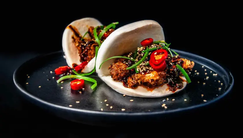 Zwei köstliche Bao Buns, gefüllt mit knusprigem Fleisch, garniert mit Chilis und Frühlingszwiebeln, auf einem schwarzen Teller.