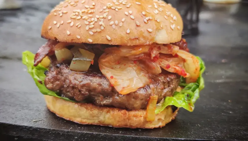 Ein saftiger korean burger mit Kimchi, Gurken und Speck auf einem Sesambrötchen.