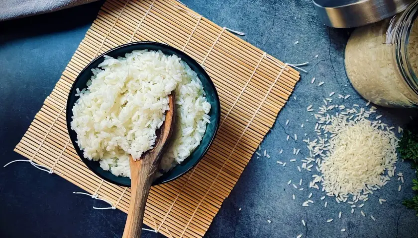 Schüssel mit gekochtem Reis auf Bambusmatte, daneben ungekochter Reis. Die Frage "wie lange muss Reis kochen" wird hier visuell beantwortet.