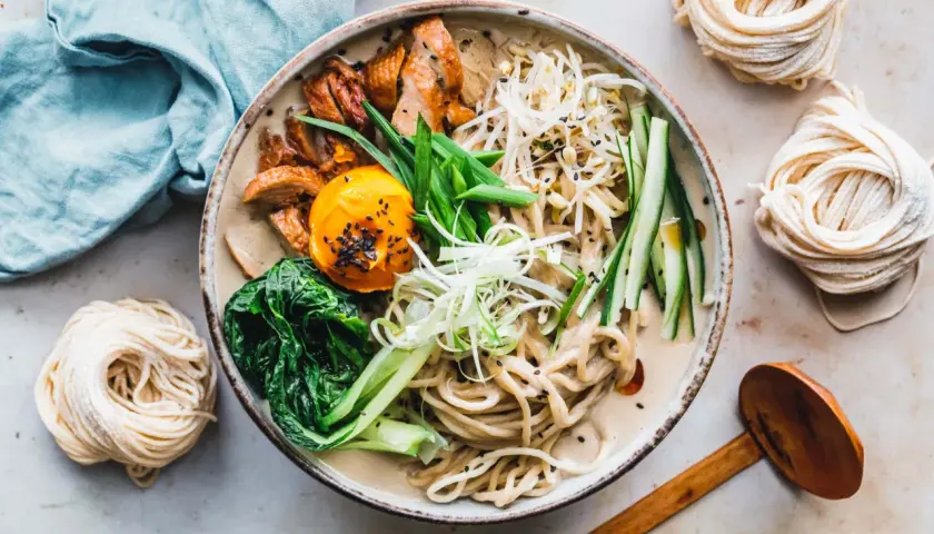 Schüssel mit cremiger Ramen-Suppe, Hühnchen, Ei, Gemüse und frischen **Rahmen Nudeln** daneben.