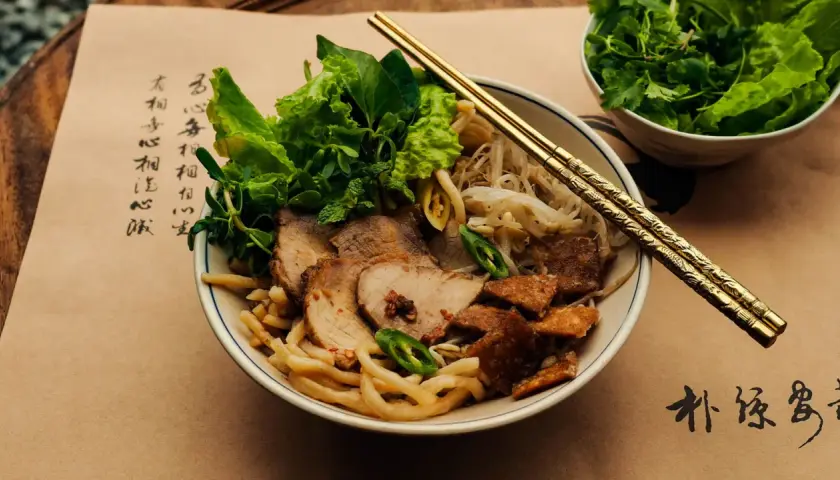 Cao Lầu: Nudelschüssel mit Schweinefleisch, Sprossen, Kräutern und knusprigen Krümeln. Goldene Stäbchen ruhen darauf.