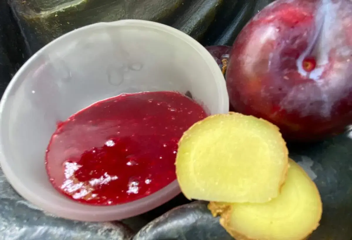 Eine Schale mit leuchtend roter asiatischer Pflaumensauce neben frischen Pflaumen und Ingwerscheiben.