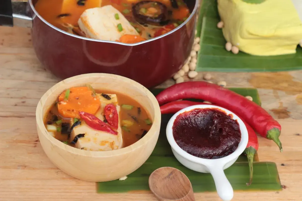 Gochujang Zutaten: Ein Topf mit Tofu-Eintopf, eine Schüssel mit scharfem Eintopf, rote Chilis, Sojabohnen und ein Block Tofu auf Bananenblättern.