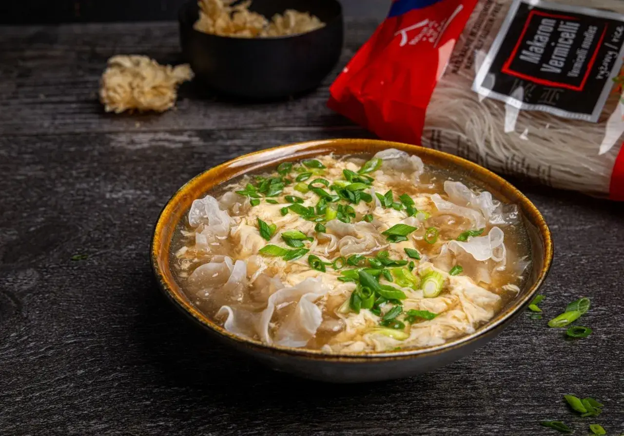 Eine dampfende Schüssel chinesische Suppe mit Ei, garniert mit Frühlingszwiebeln und Pilzen. Daneben liegt eine Packung Reisnudeln.
