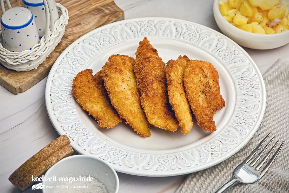 Knusprige Hähnchenstreifen in goldbrauner Panko Panade auf einem Teller, daneben Kartoffelsalat.