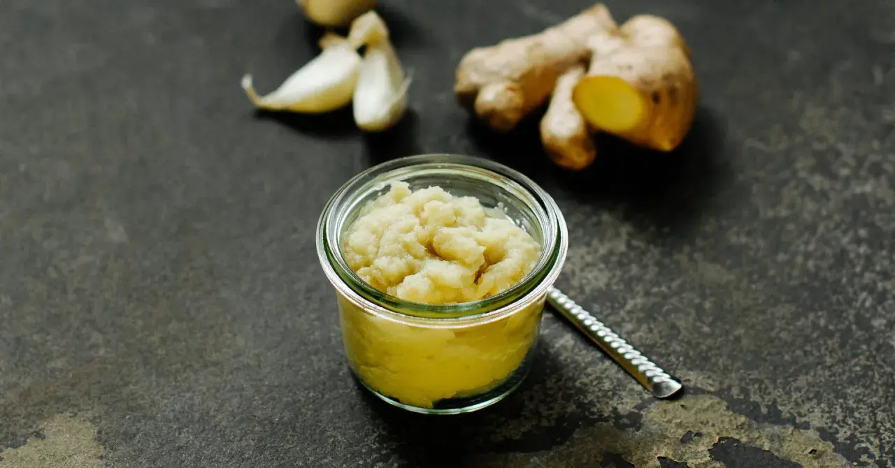 Frisch zubereitete ingwer knoblauch paste in einem Glas. Daneben liegen frische Knoblauchzehen und Ingwerwurzeln.