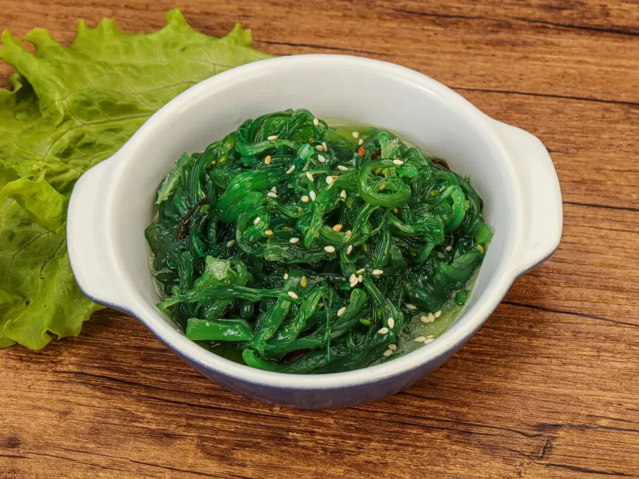 Schüssel mit leuchtend grünem Seetang Salat, garniert mit Sesam. Daneben ein Blatt grüner Salat.