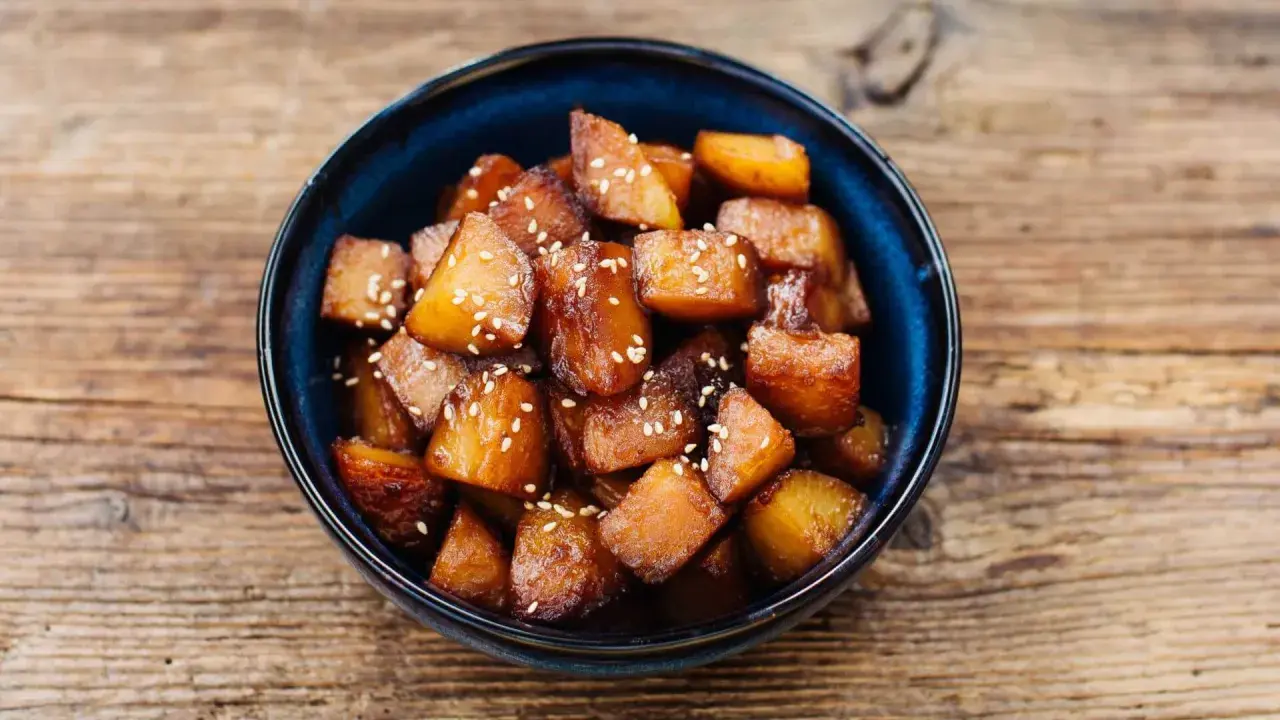 Glasierte Süßkartoffelwürfel mit Sesam, ein köstliches Beispiel für Banchan Rezepte, serviert in einer blauen Schüssel auf Holzuntergrund.