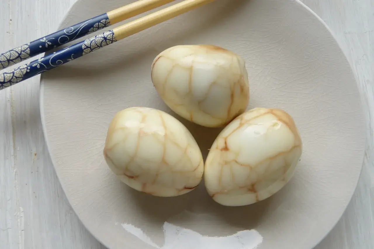 Drei marmorierte Tee Eier auf einer hellen Platte, daneben blaue Essstäbchen.