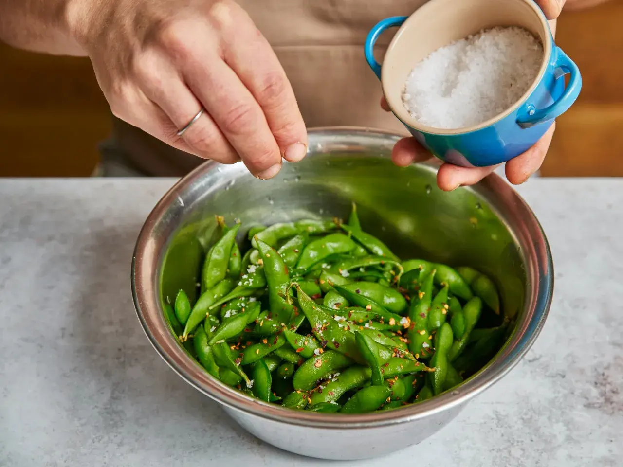 Hände würzen frisch gekochte Edamame mit Meersalz und Chiliflocken. So einfach ist es, Edamame zuzubereiten!