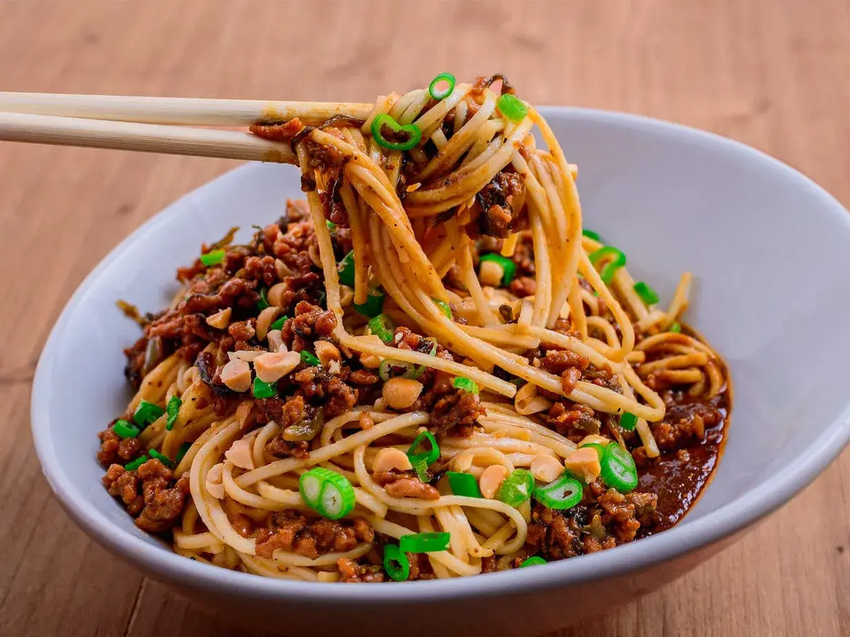 Ein Teller Dan Dan Noodles mit Hackfleisch, Erdnüssen und Frühlingszwiebeln, serviert mit Stäbchen.