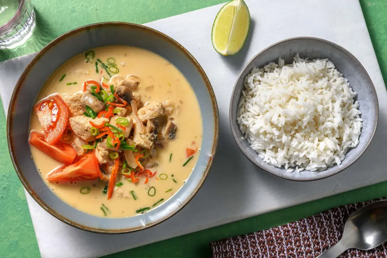 Khao tom: Eine Schüssel mit cremigem Hühner-Kokos-Eintopf, garniert mit Tomaten und Frühlingszwiebeln, daneben eine Schüssel Reis und eine Limettenspalte.