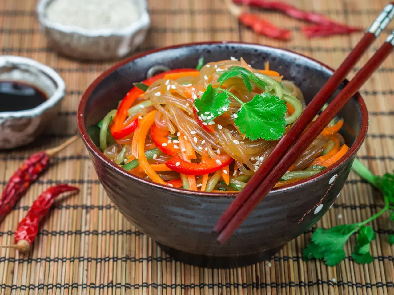 Schüssel mit Japchae, einem koreanischen Nudelgericht, garniert mit Koriander und Sesam. Daneben Sojasauce, getrocknete Chilis und Reis.