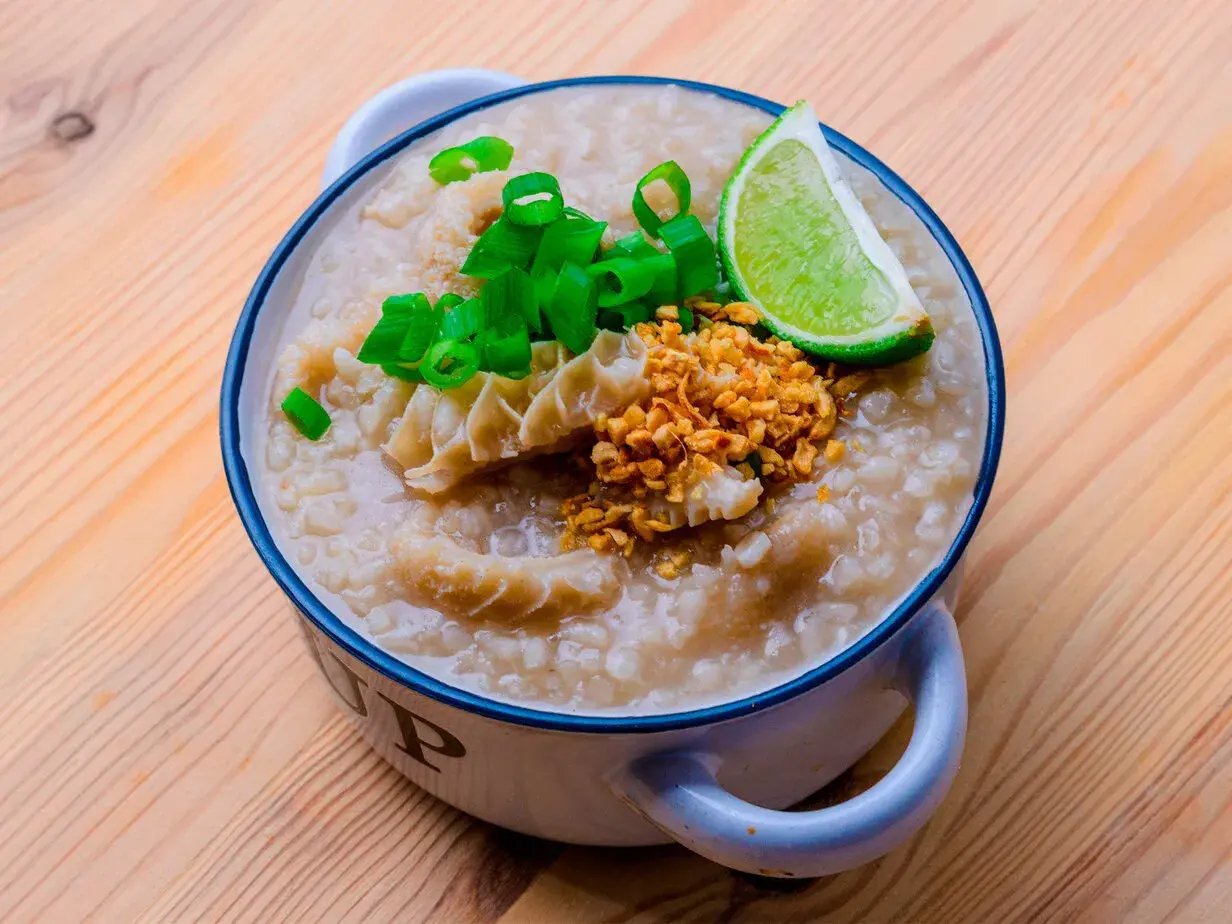 Herzhafter Reis Congee mit Rinderpansen, Frühlingszwiebeln, geröstetem Knoblauch und Limettenspalte in einer Schüssel auf Holzhintergrund.