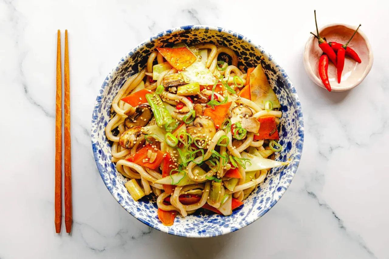 Schüssel mit leckeren Udon Nudeln kochen, garniert mit Gemüse, Frühlingszwiebeln und Sesam. Daneben liegen Essstäbchen und eine kleine Schale mit Chilis.