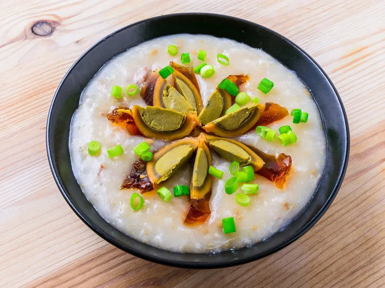 Ein cremiges Congee-Rezept mit geschnittenem konserviertem Ei und Frühlingszwiebeln, serviert in einer schwarzen Schüssel auf Holzhintergrund.