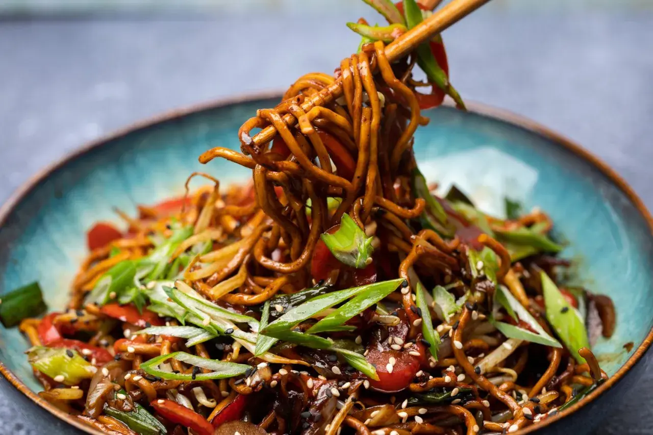 Ein Teller mit dampfendem Chow Mein, garniert mit Frühlingszwiebeln und Sesam. Essstäbchen heben eine Portion der Nudeln an.