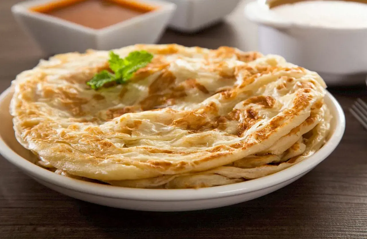 Ein Stapel knuspriger Roti Canai, garniert mit Minze, serviert auf einem Teller mit Curry und Joghurt im Hintergrund.