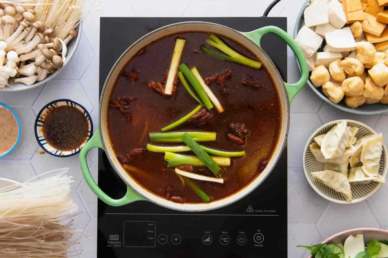 Ein köstlicher chinesischer Eintopf brodelt in einem grünen Topf, umgeben von Zutaten wie Pilzen, Tofu, Teigtaschen und Reisnudeln.