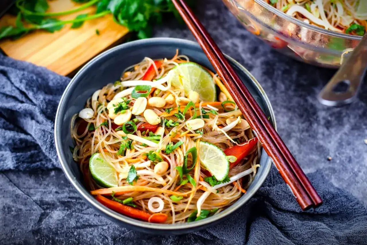 Schüssel mit asiatischem Glasnudelsalat, garniert mit Erdnüssen, Limettenspalten und Frühlingszwiebeln. Chopsticks liegen auf dem Teller.
