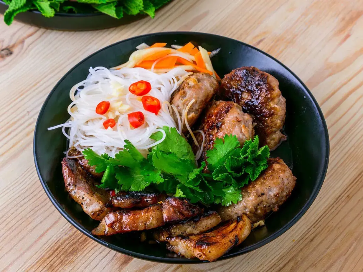 Bun cha Hanoi: gegrilltes Schweinefleisch und Fleischbällchen mit Reisnudeln, Kräutern und Chili auf einem Teller.
