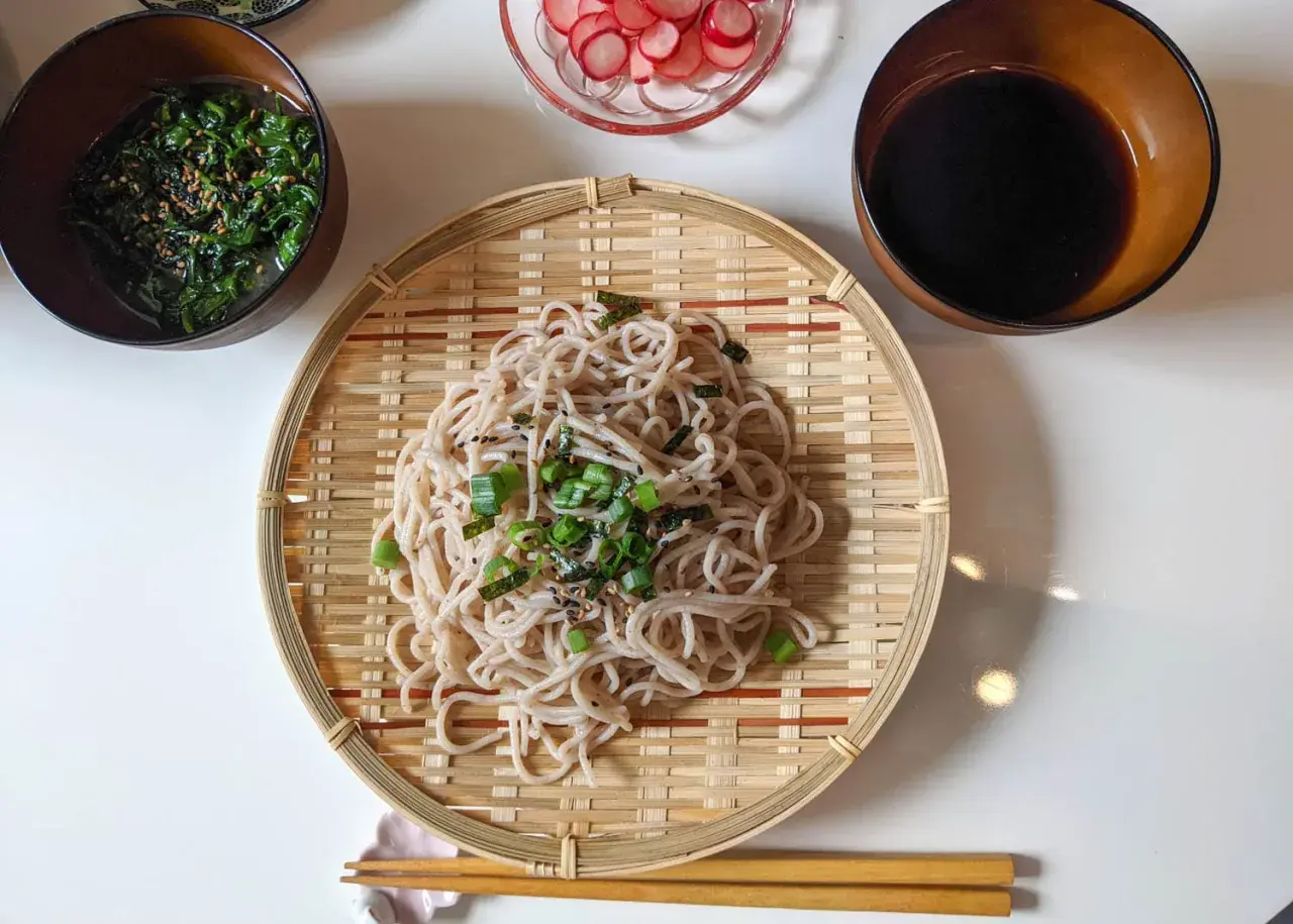 Kalte Soba-Nudeln, garniert mit Frühlingszwiebeln und Nori, serviert auf einem Bambusteller mit Beilagen wie Seetangsalat und Radieschen.