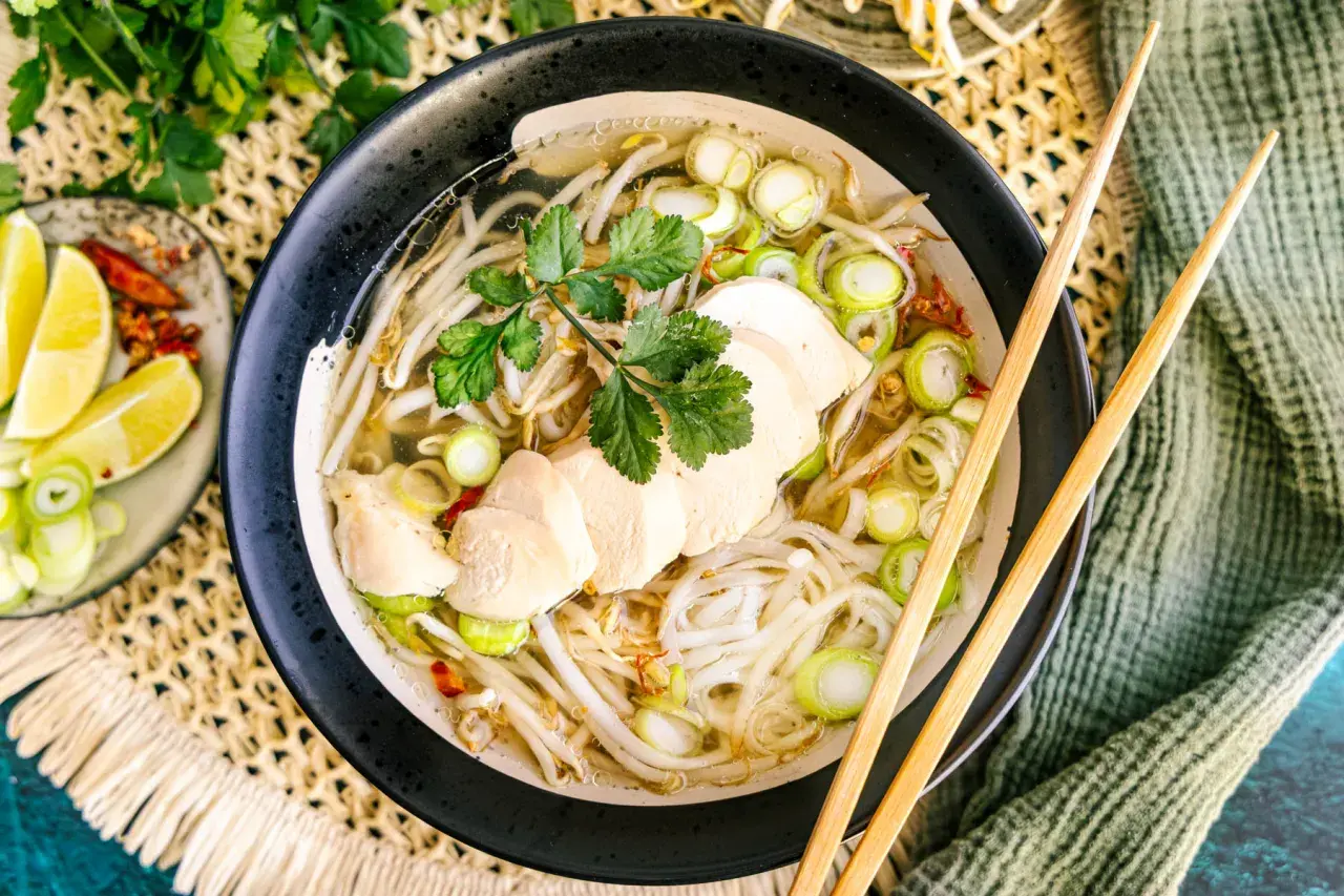 Eine dampfende Schüssel Pho Ga mit zarten Hühnerbrustscheiben, Reisnudeln, Sojasprossen und Frühlingszwiebeln. Dazu Limettenspalten und Chili.