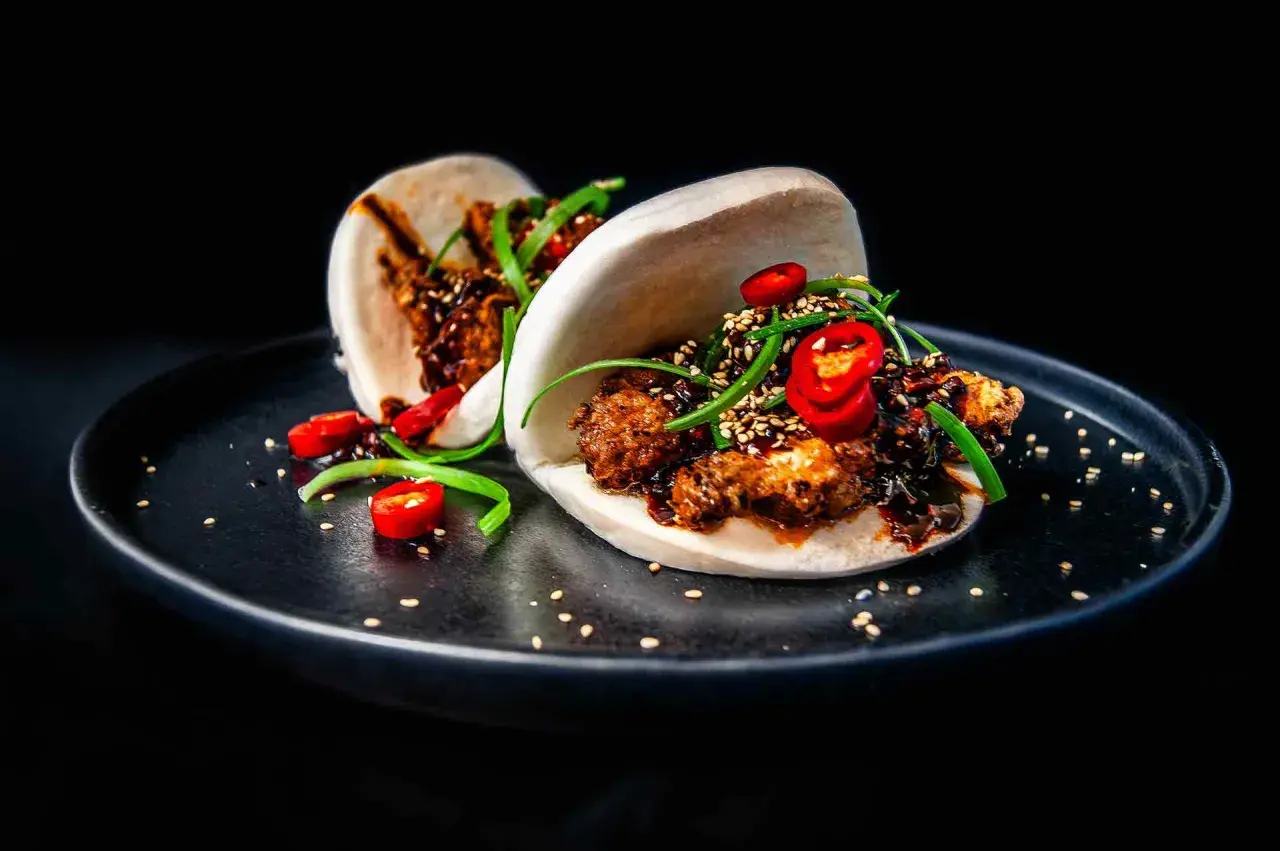 Zwei köstliche Bao Buns, gefüllt mit knusprigem Fleisch, garniert mit Chilis und Frühlingszwiebeln, auf einem schwarzen Teller.